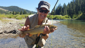 woman smiling with a fish with a lake, trees, and mountain in the background