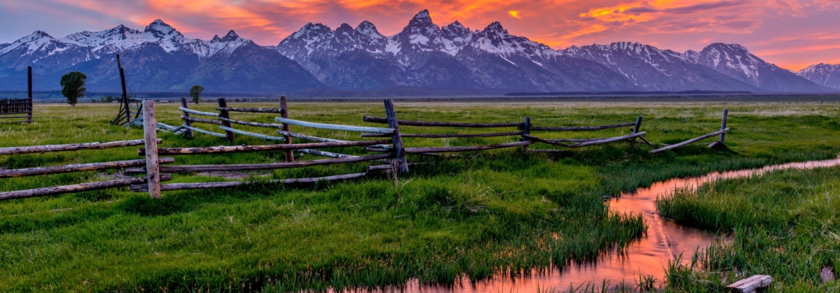A sunset over the Grand Teton mountains with a rustic fence and open field in the foreground.