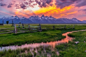A sunset over the Grand Teton mountains with a rustic fence and open field in the foreground.