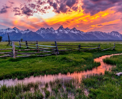 A sunset over the Grand Teton mountains with a rustic fence and open field in the foreground.